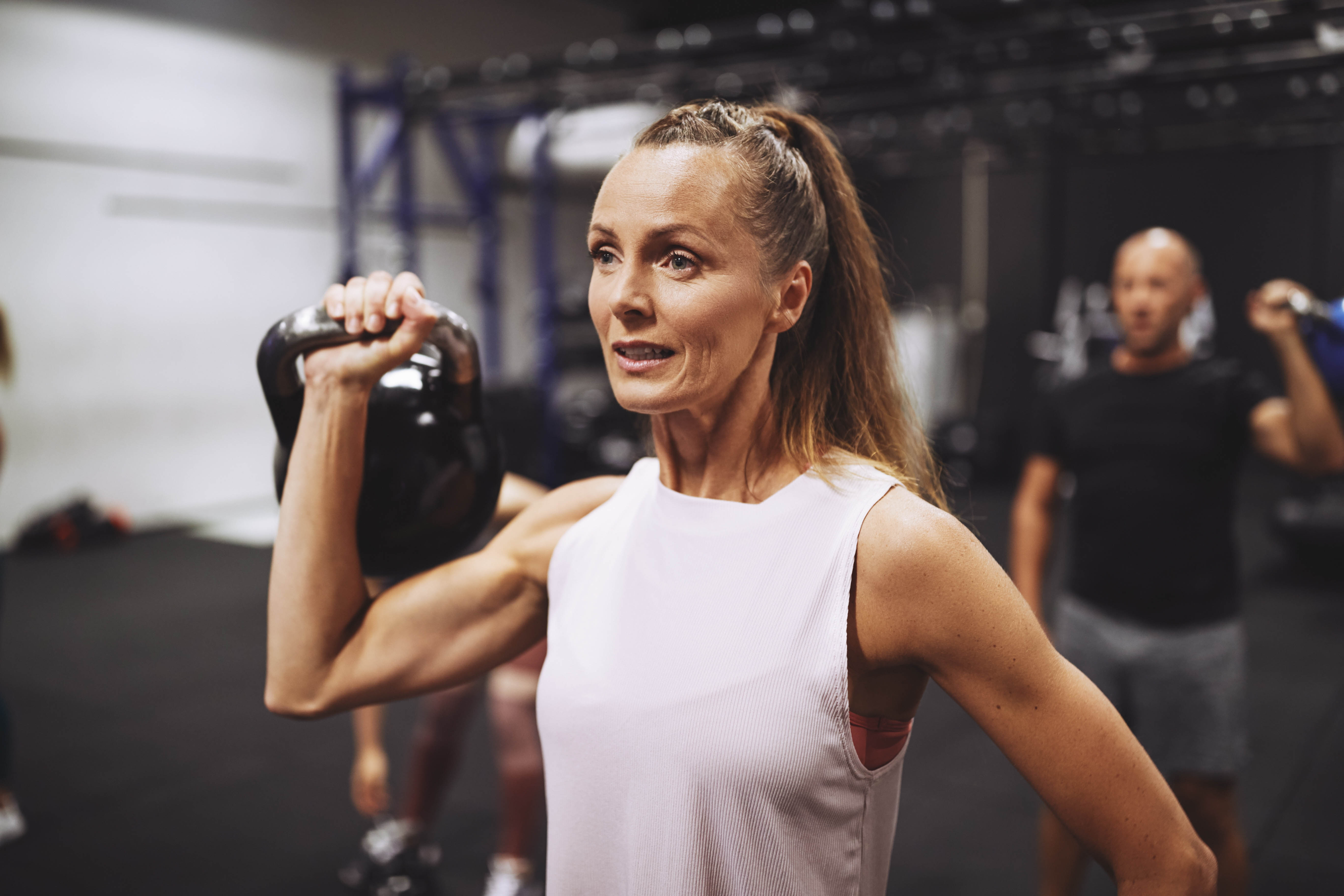 Strong, middle aged woman standing in a gym mid-workout, with a kettlebell in her hand at her shoulder height Strong, middle aged woman standing in a gym mid-workout, with a kettlebell in her hand at her shoulder height